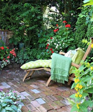 A backyard area with a brick patio with a wooden sunlounger with lime green cushions and different sized green bushes and trees around it with red, white, and yellow flowers