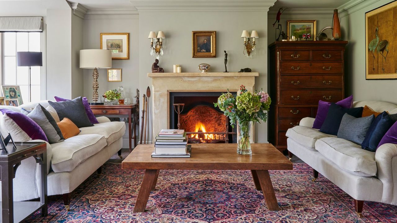 An eclectic living room with a large marble lit fireplace, patterned purple rug, light matching sofas with assorted pillows, a large wooden coffee table with stacks of books and a vase of large flowers, a dark brown chest of drawers, framed artworks and sage green walls