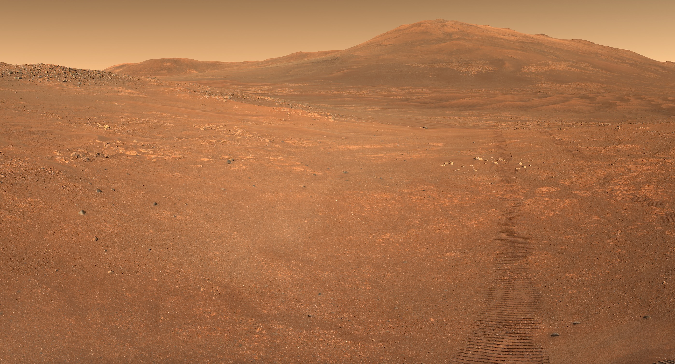 foto de Marte tirada por um veículo espacial, mostrando um deserto de terra vermelha com uma montanha ao fundo e uma linha de rastros do veículo espacial em primeiro plano