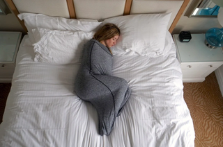 A woman lying in a bed with white sheets inside a grey swaddle blanket pod