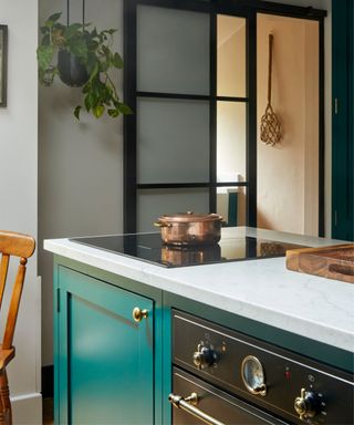 A stylish kitchen island with teal cabinetry, a marble countertop, and brass hardware. The island features an integrated induction hob with a copper pot on top. A black-framed sliding door with frosted glass adds a modern touch, while a hanging plant and natural textures enhance the space.