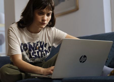 A young woman with dark hair sits on a blue couch, focused on a silver HP laptop in front of her, wearing a white Coney Island graphic T-shirt.