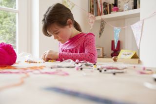 A young brunette girl sat at a table and making something, surrounded by crafting materials such as wool and card.
