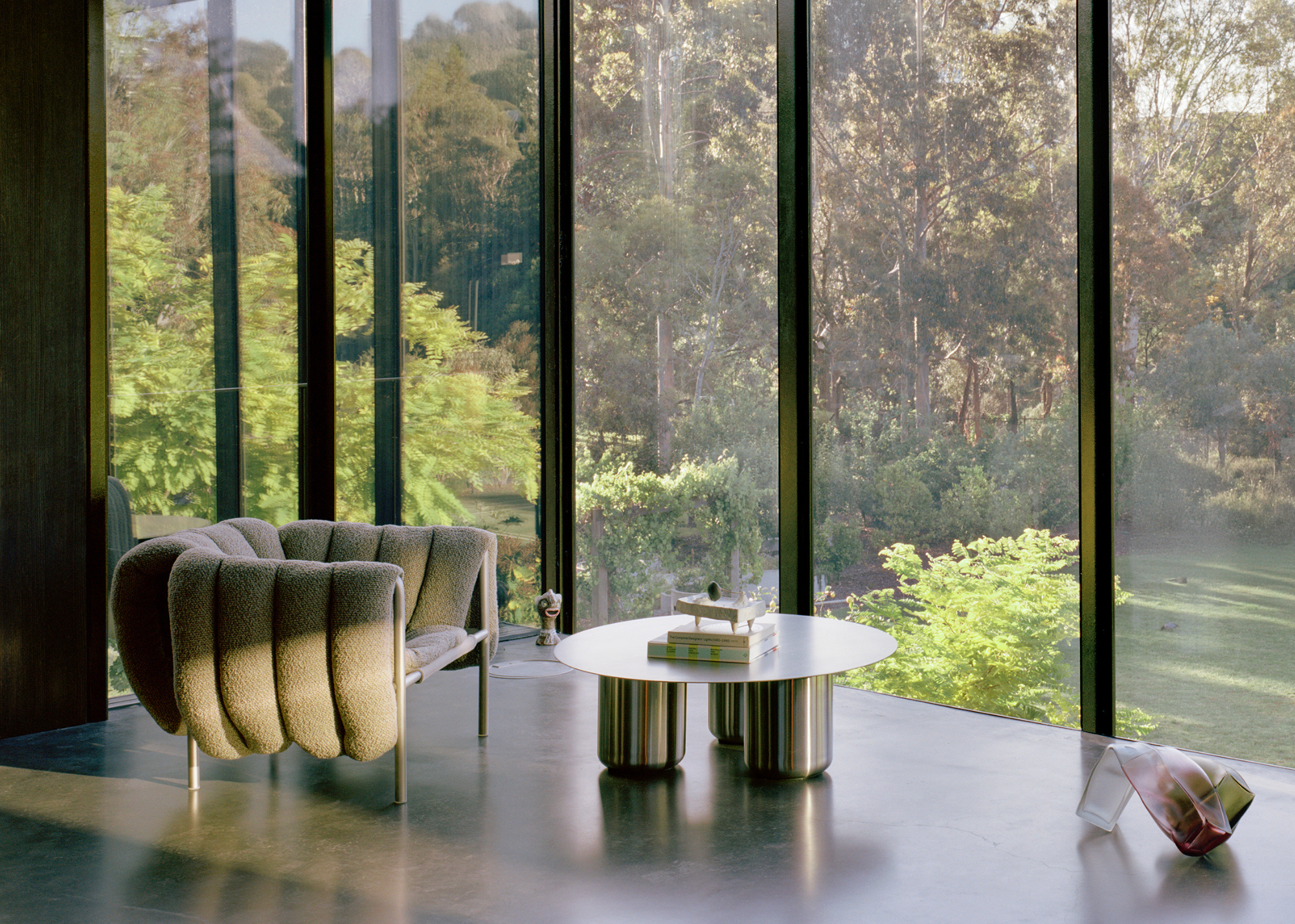 A living space overlooking a lush backyard through glass windows with a green armchair, a steel coffee table, and a structural glass piece