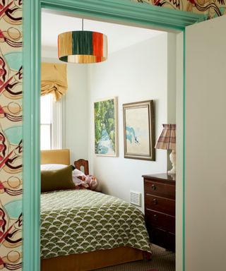 patterned hall wallpaper looking into bedroom with yellow headboard, green patterned throw and colorful pendant light
