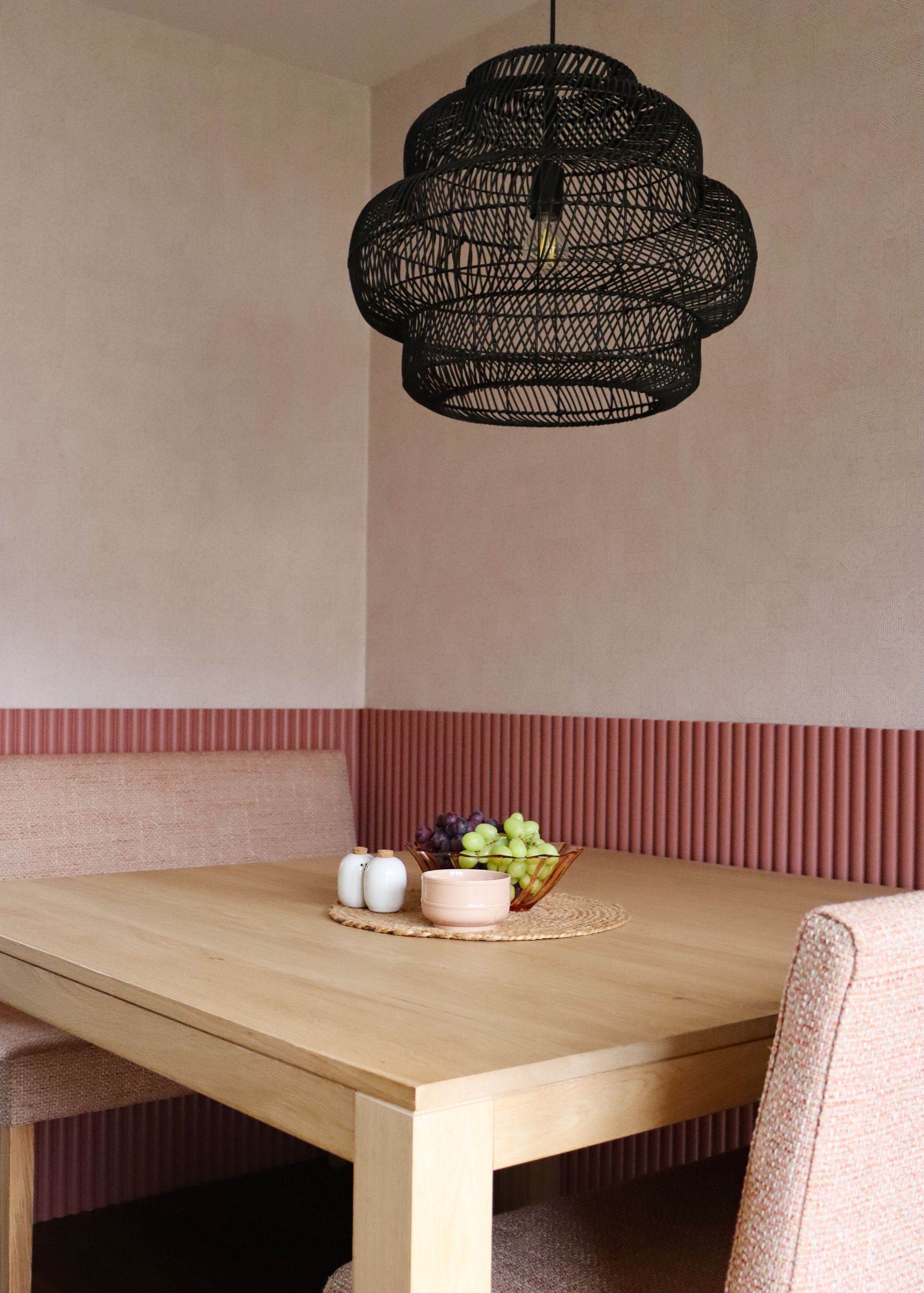 A dining table with baby pink dining chairs. On top of the table is a bowl of grapes, and salt and pepper shakers. Above the table is a boho themed pendant light