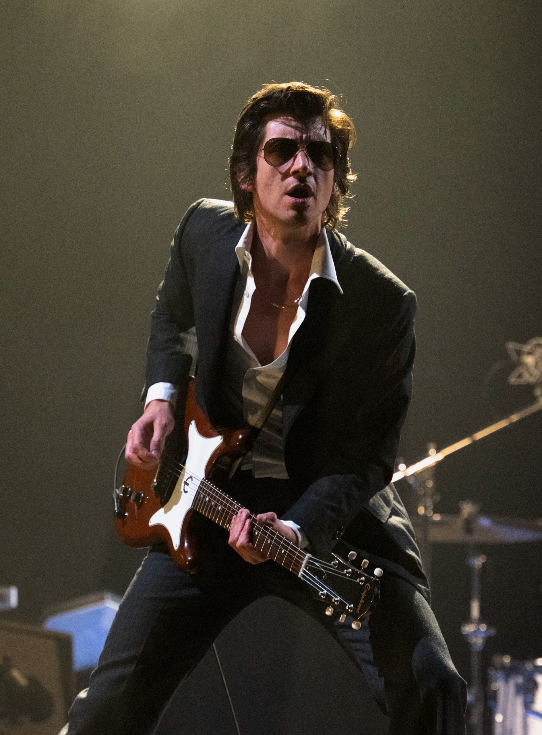 Alex Turner plays a sunburst electric guitar on stage in an open shirt and aviator sunglasses, shot from a low angle against a dark arena backdrop.