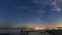 multiple meteors streak through a star studded sky with the milky way off to the right. there are lights shining over the water in the distance.