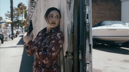 a woman (Juliana Aidén Martinez as June Hawkins) hides behind a wall while holding a gun