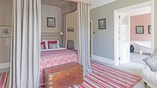 A grey bedroom with four-poster bed with curtains, a red and cream striped rug, wooden trunk, and open doors to a peachy pink ensuite to the right with a rolltop bath