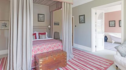 A grey bedroom with four-poster bed with curtains, a red and cream striped rug, wooden trunk, and open doors to a peachy pink ensuite to the right with a rolltop bath
