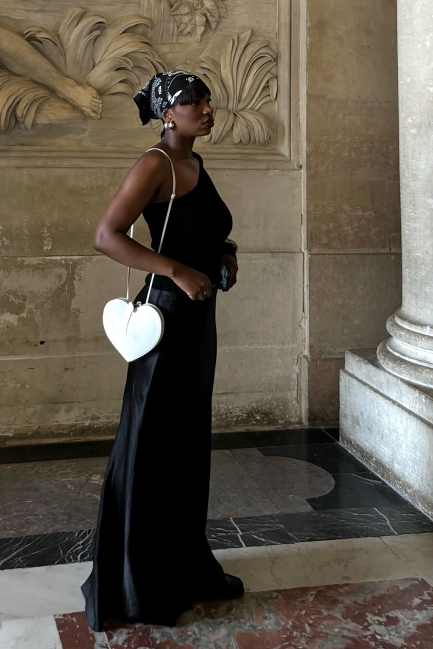 How to wear a bandana is shown in a photo of a woman standing in a marble foyer wearing a black bandana, silver hoops, black asymmetrical top, black satin maxi skirt, white shoulder bag, and black flats