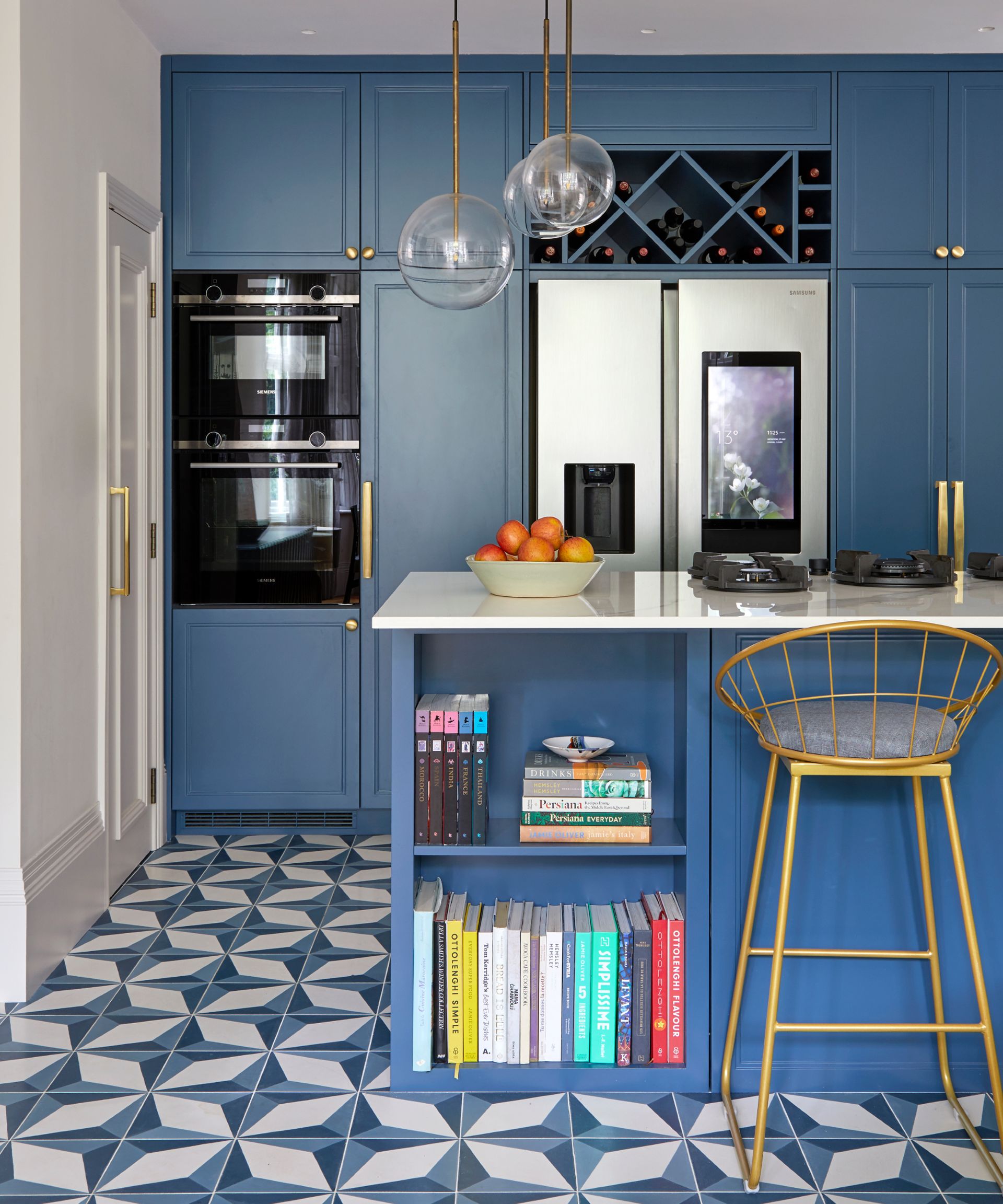 mid blue kitchen with built in double oven and refrigerator, wine storage above. Brass knobs and handles, brass bar stool, blue and cream 3d style patterned floor tiles, white countertop on island, trio of glass pendants