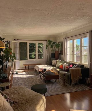 A small living room with a white ceiling, windows, gray couch, rug, and footstools and poufs