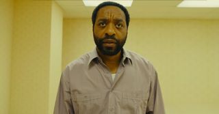 Chiwetel Ejiofor standing in the Backrooms, where he is surrounded by pale yellow walls and fluorescent lights.