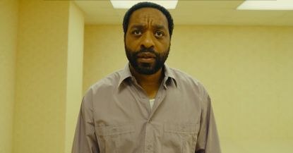 Chiwetel Ejiofor standing in the Backrooms, where he is surrounded by pale yellow walls and fluorescent lights.