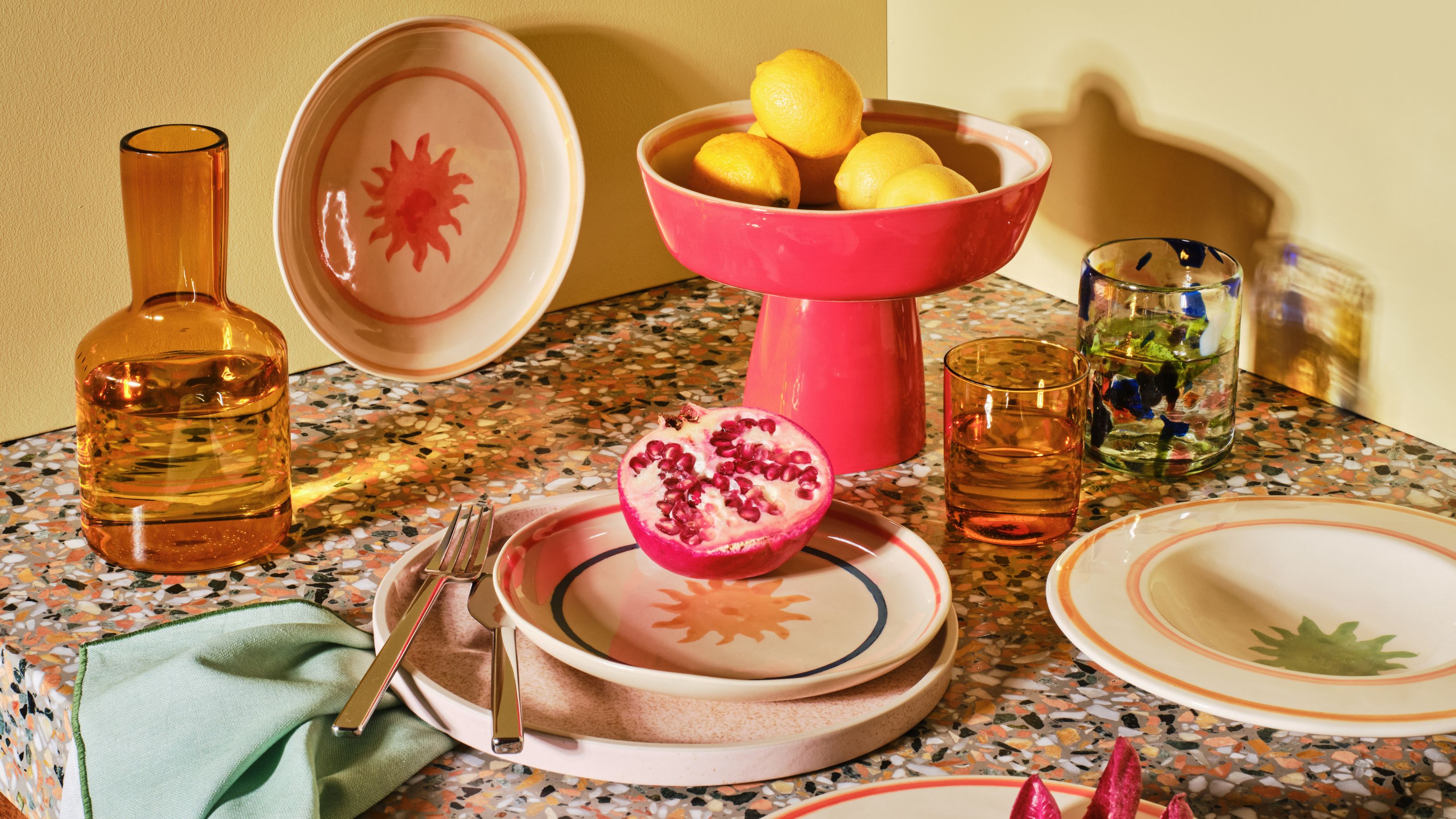 homewares, including an orange glass carafe, colorful dinner plates, a pink plinth bowl filled with lemons, a green napkin, colorful glassware and cut pomegranate, styled on a terrazzo countertop