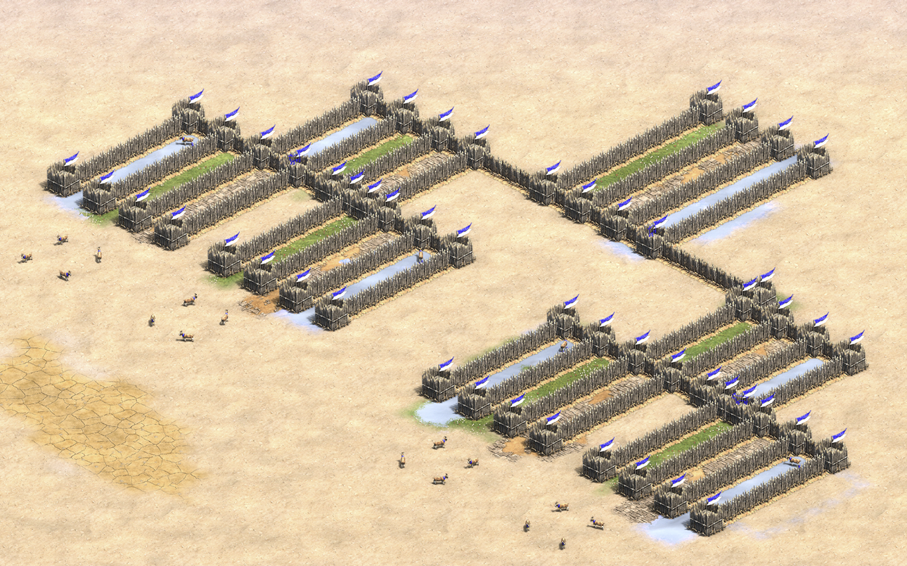 An aerial view of an Age of Empires 2 wooden palisade settlement layout with green fields and blue flags on a sandy background, featuring small figures moving in the foreground.
