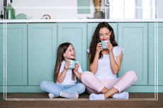 Dreamy mother sitting on kitchen floor together with daughter, drinking coffee, enjoying morning beverage and spending weekend in cozy apartment, resting at home.