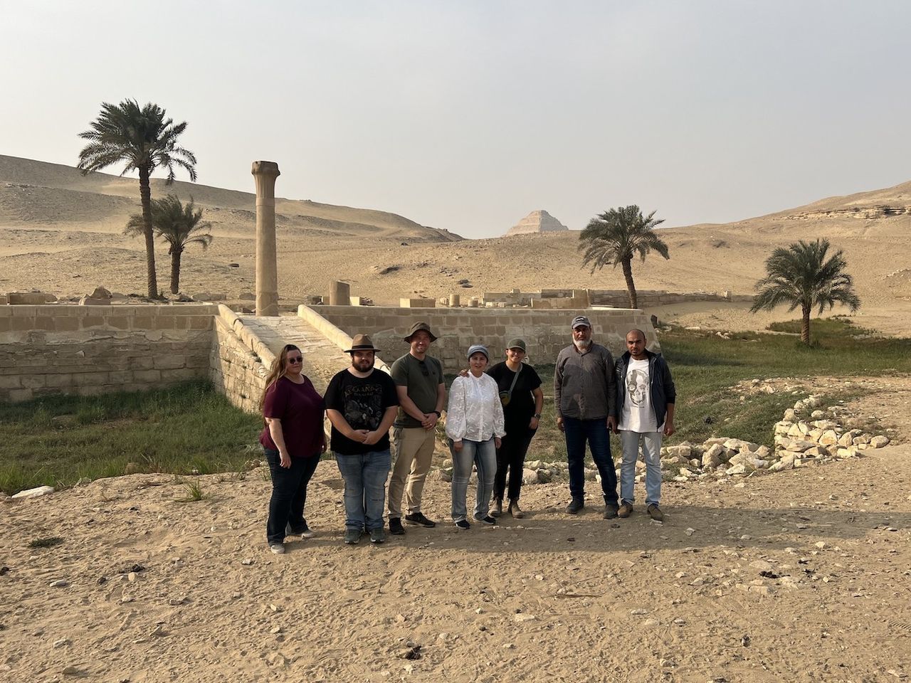 A team of seven people stands in front of a stone base with a pillar on top of it. A few palm trees are also in the background and a pyramid