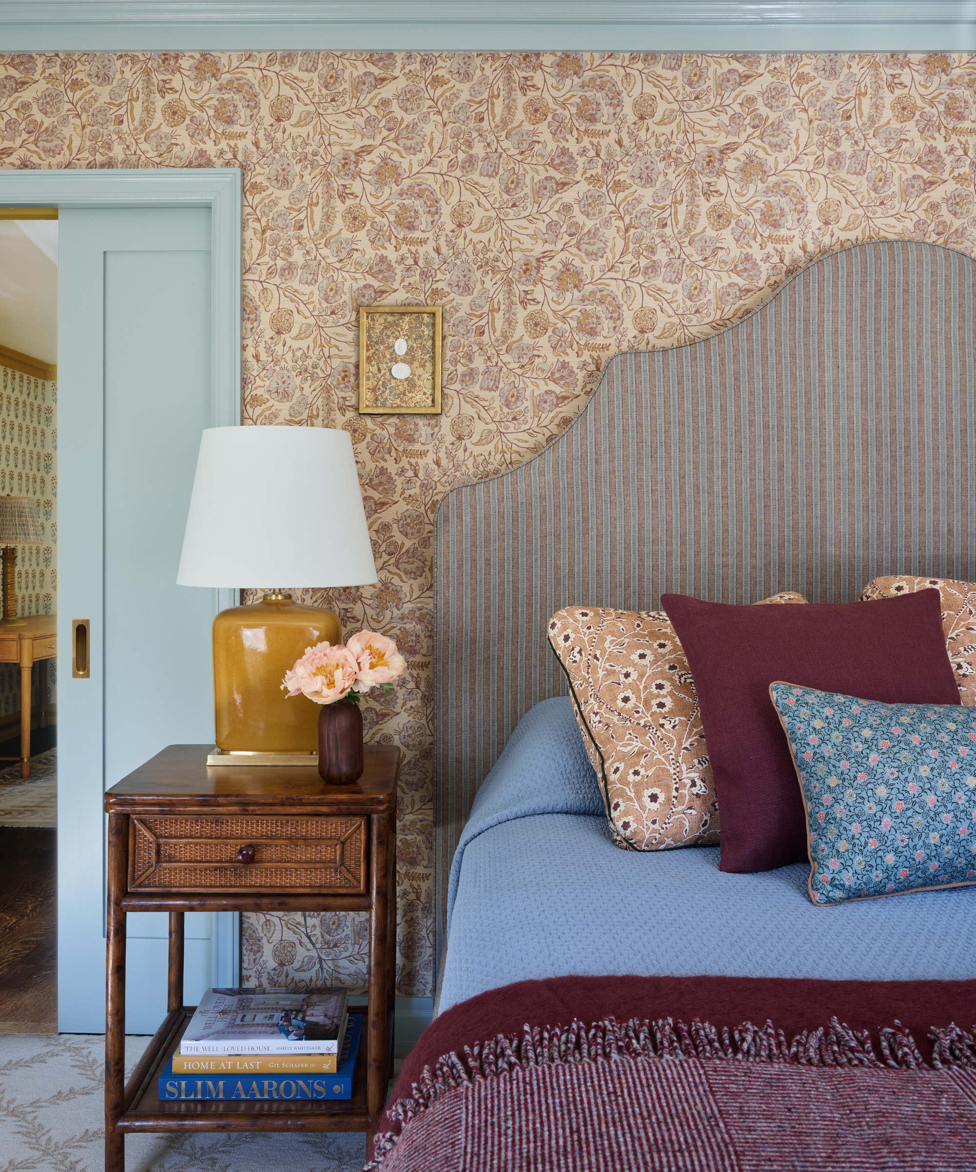Primary bedroom with floral wallpaper, striped upholstered headboard, cane nightstand and soft blue trim.