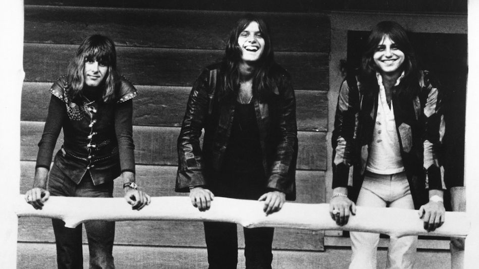 Keith Emerson, Carl Palmer and Greg Lake standing on the porch of a wooden cabin