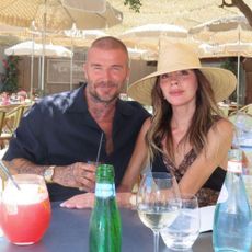 Victoria Beckham sits next to David Beckham wearing a lace trim black dress with an oversized straw hat.