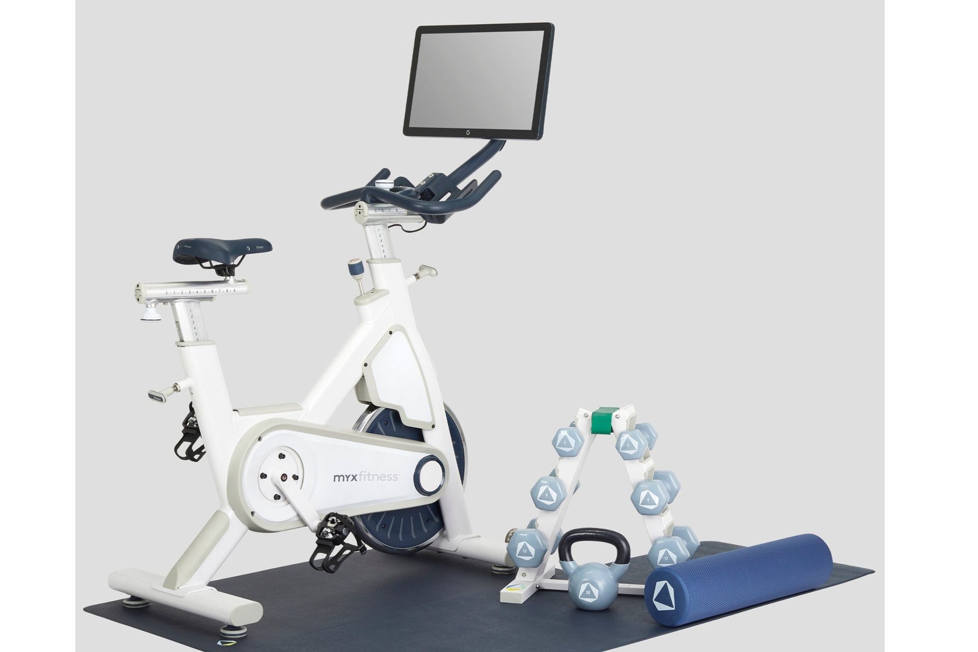 MYXFitness MYX II Plus is show in the image on the left, with it's integrated screen above the handlebars of the bike in the top middle. The bike is on a matt and next to it on the right are dumbbells on a rack, a kettlebell and a foam roller