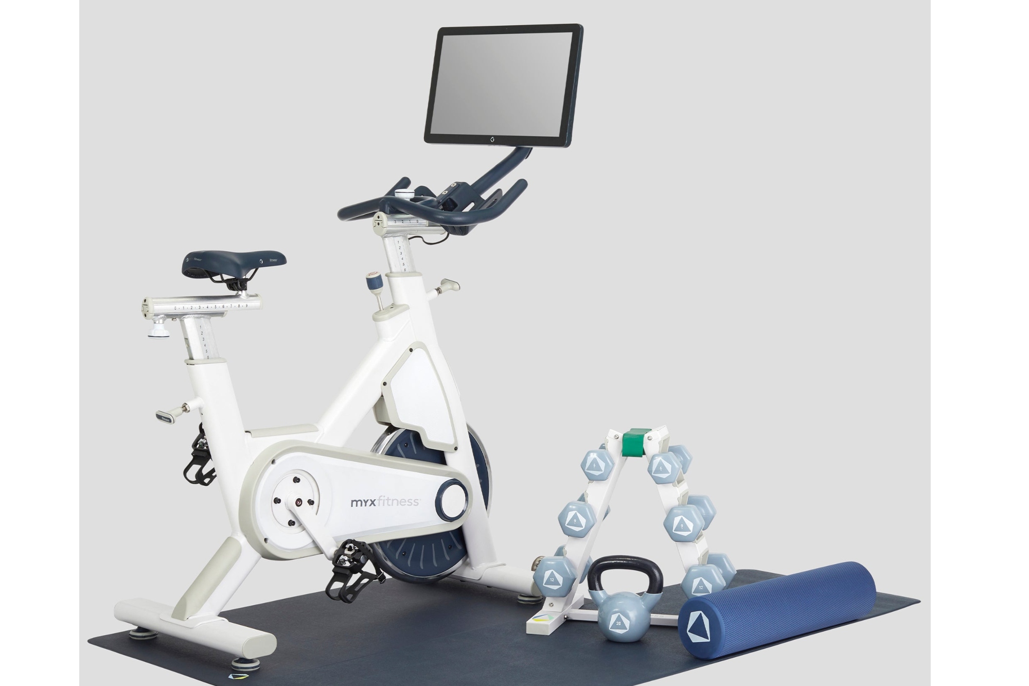 MYXFitness MYX II Plus  with it's integrated screen above the handlebars of the bike in the top middle. The bike is on a matt and next to it on the right are dumbbells on a rack, a kettlebell and a foam roller