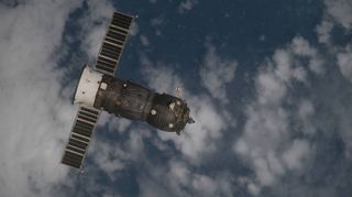 A Russian Progress spacecraft with solar panels extended flies in orbit above an ocean partially covered by clouds.