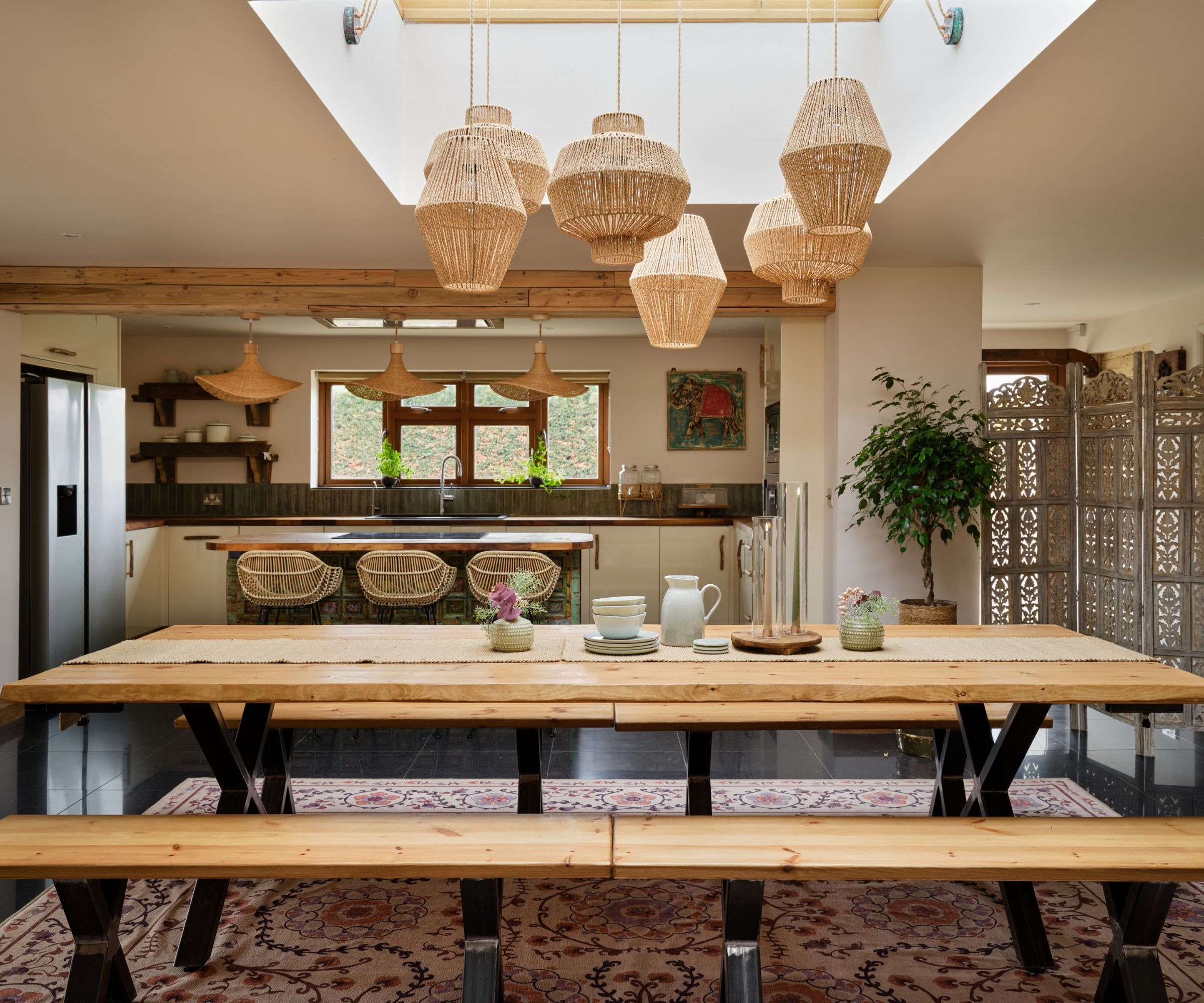 An open plan kitchen and dining area with a large wooden picnic-style table with a rattan pendant light above and candles in hurricane glasses