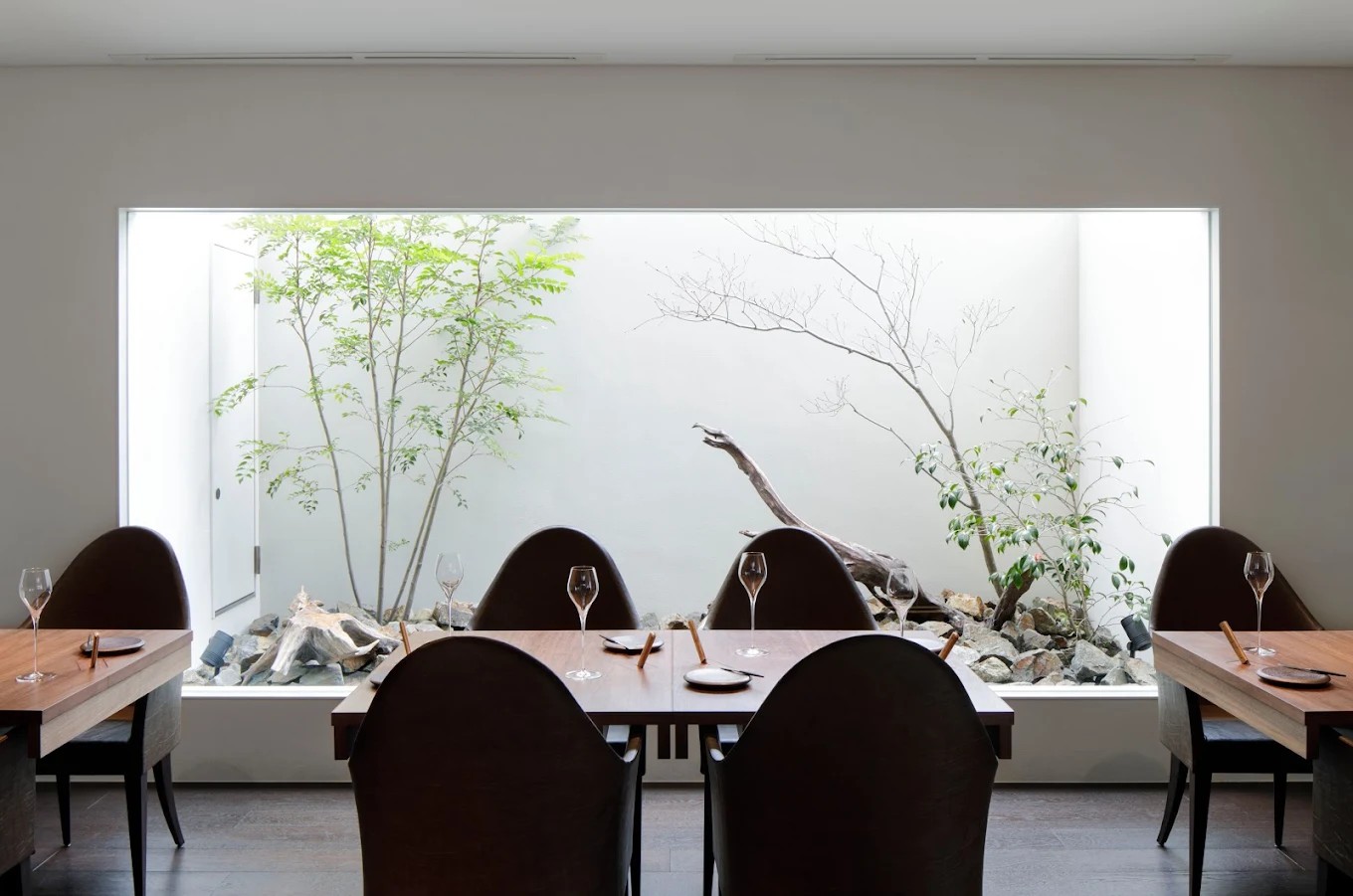 A minimally decorated restaurant with wooden tables and leather chairs features a box overlooking the outdoors, filled with plants.