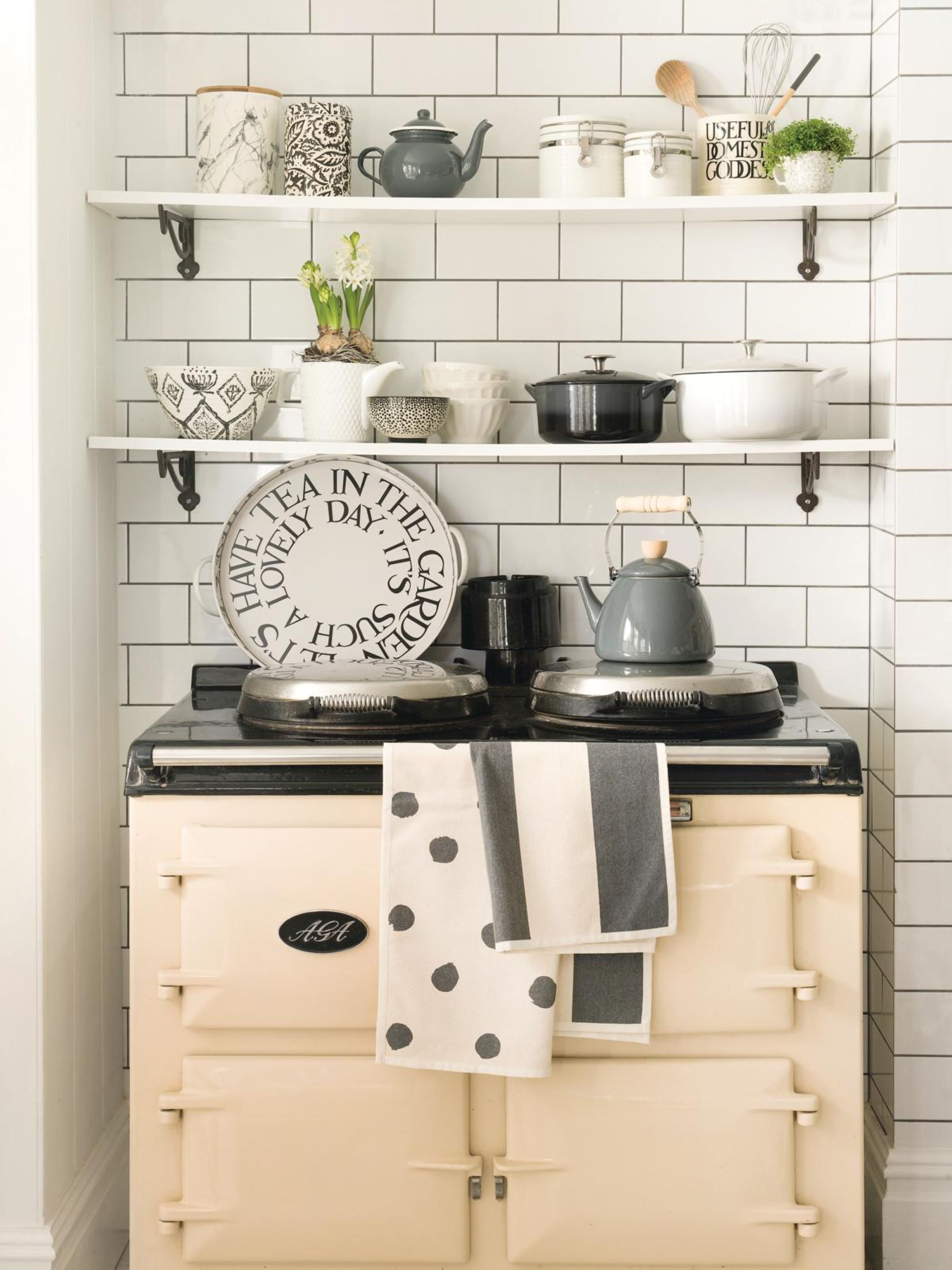 a kitchen corner with a traditional oven, two floating shelves holding kitchen objects such as pots, cups, mugs, and plants, and white tile behind