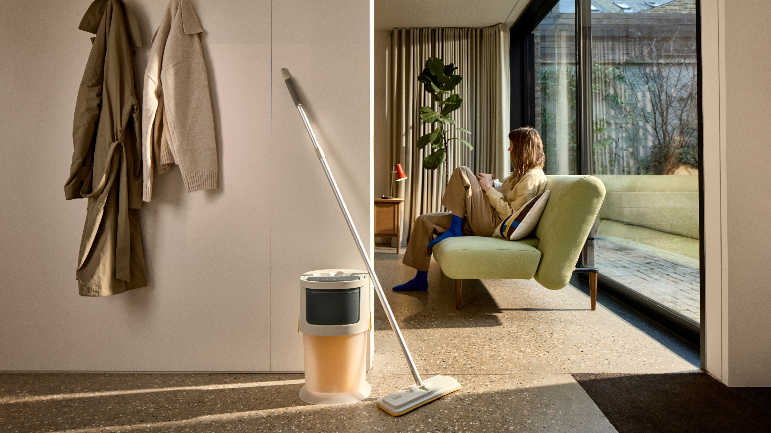 A Joseph Joseph UltraClean mop in a living room with a woman lounging on a green sofa in the back