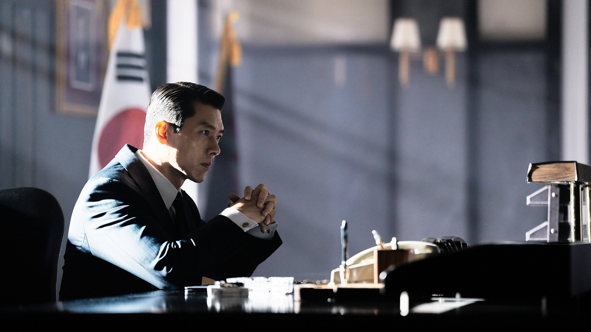 A man in a black suit (Hyun Bin as Baek Kitae) sits at an ornate desk, with the Korean flag behind him, in Hulu's 'Made in Korea.'