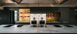 G Mimo London Cookery School Interior Featuring Caesarstone Rugged Concrete Worktops, grey kitchen cupboards, open shelves and stainless steel ovens