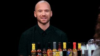 Sean Evans smiles while sitting in front of Season 25's line of hot sauces for Hot Ones.