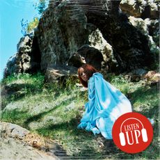 singer ravyn lenae wearing a blue dress looks over her shoulder while climbing up a mountain on the cover of her album blue island with the marie claire listen up franchise stamp over it