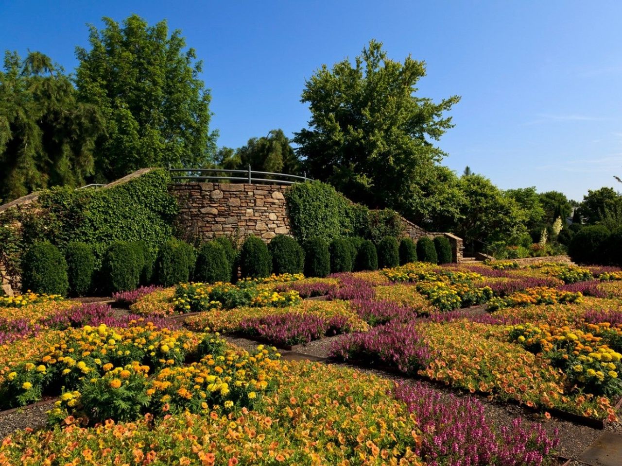 A beautiful patterned garden in shades of purple and orange overlooked by a stone wall and trees