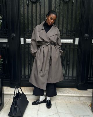 Woman wearing grey trench coat, black pants, black turtleneck and black loafers standing in front of gated door.