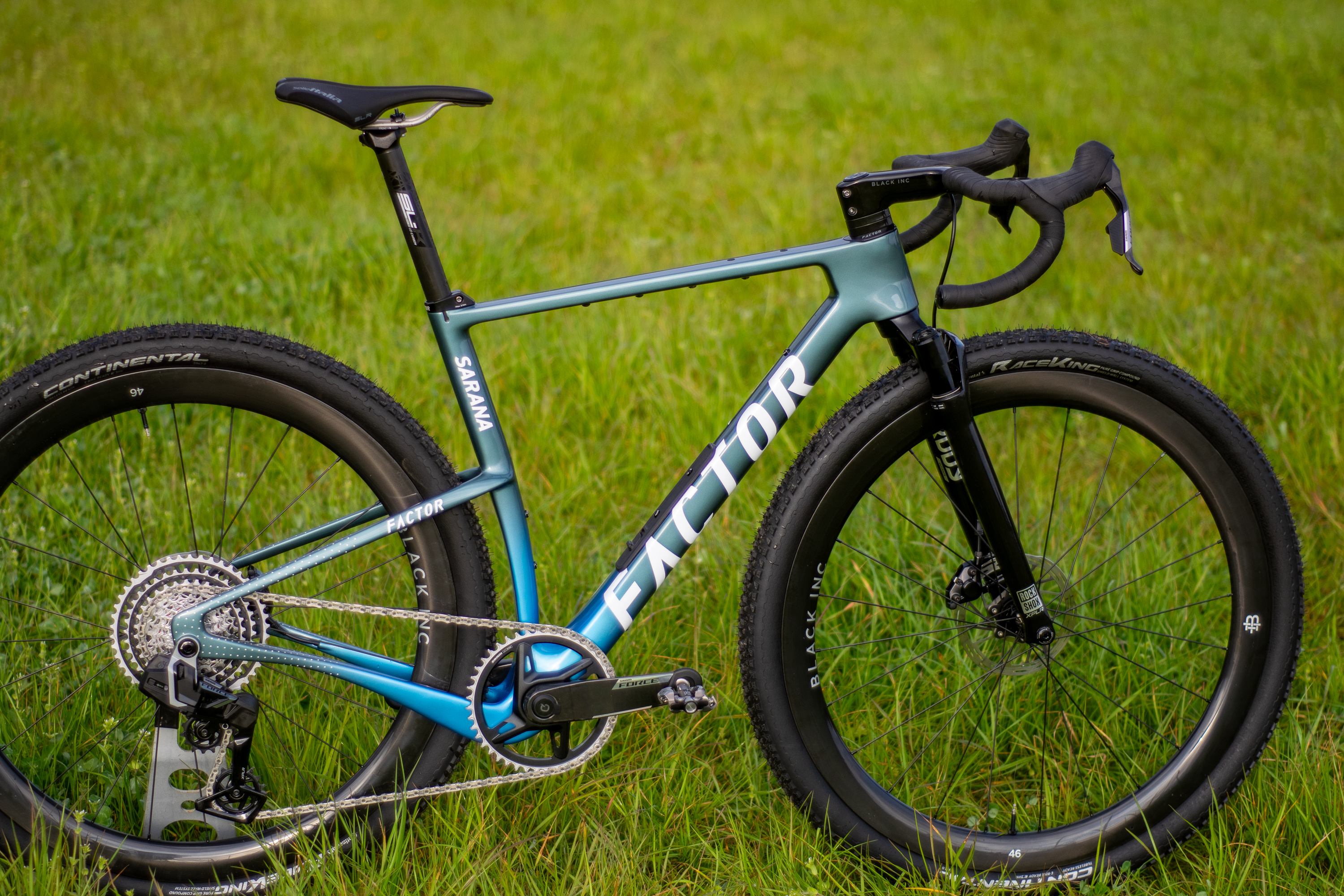 A blue Factor Sarana gravel bike in a grassy field with a hedge in the background