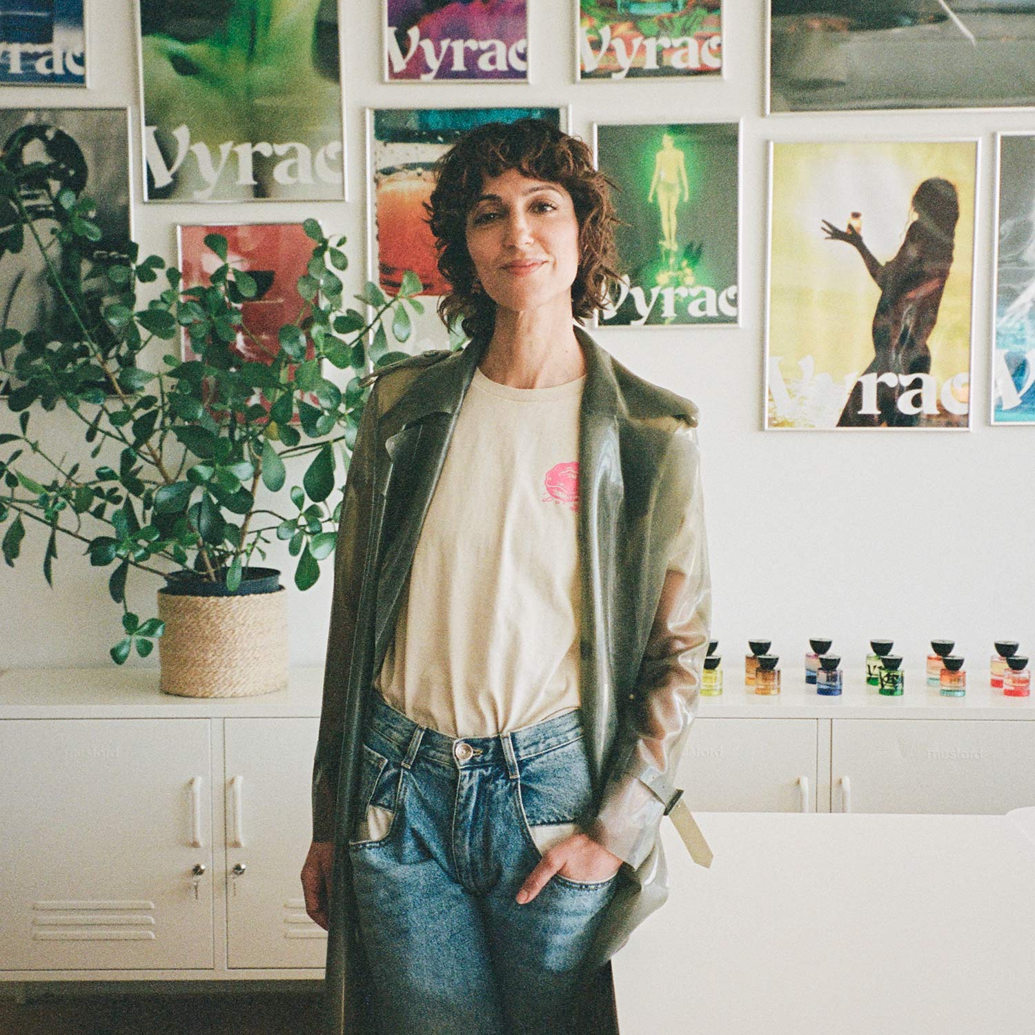 Yasmin Sewell poses in front of wall filled with posters featuring her brands Vyrao's colourful campaigns and a row of stylish perfume bottles