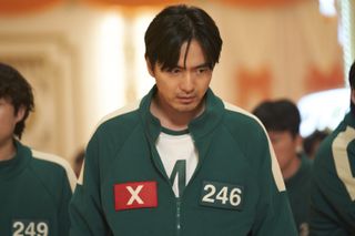 A man (Lee Jin-uk) wears a green tracksuit, with a red X patch on one side of the chest and the number 246 on the other, in 'Squid Game' season 2.