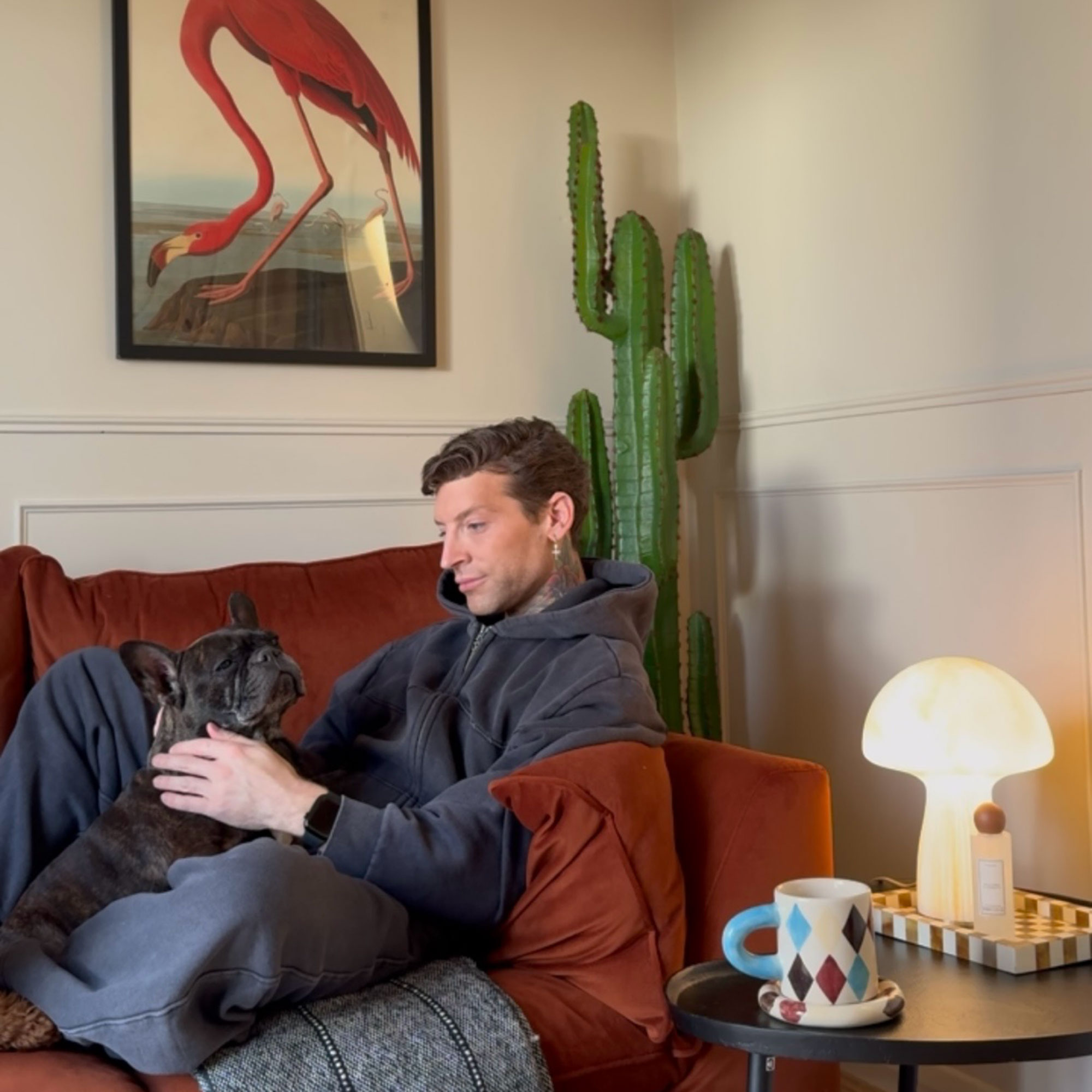Man sitting with dog on rust coloured sofa in front of cactus and flamingo print