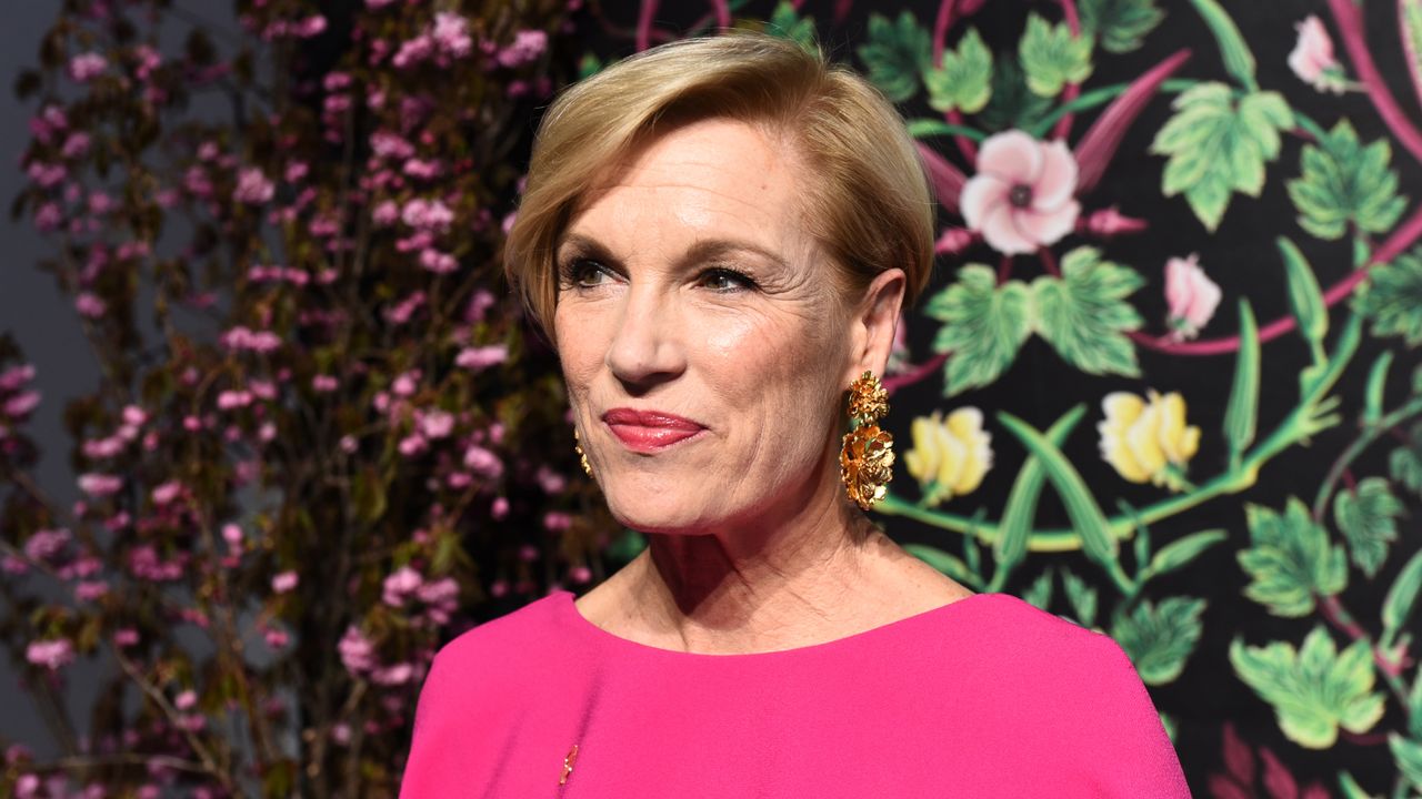 Cecile Richards wears a bright pink dress and stands in front of a wall of flowers