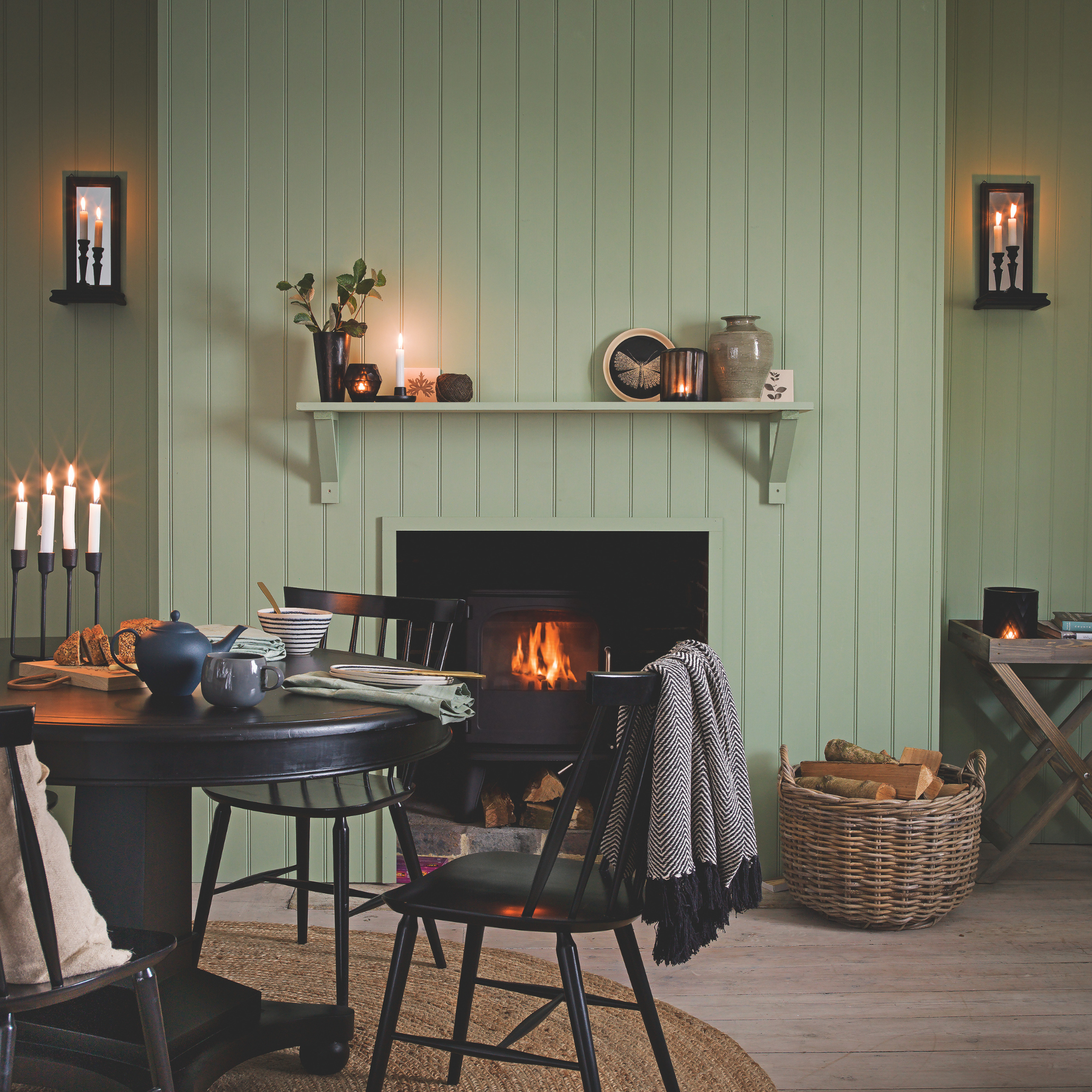 Cosy green panelled dining room with lit log burner, black dining table and chairs and candles.