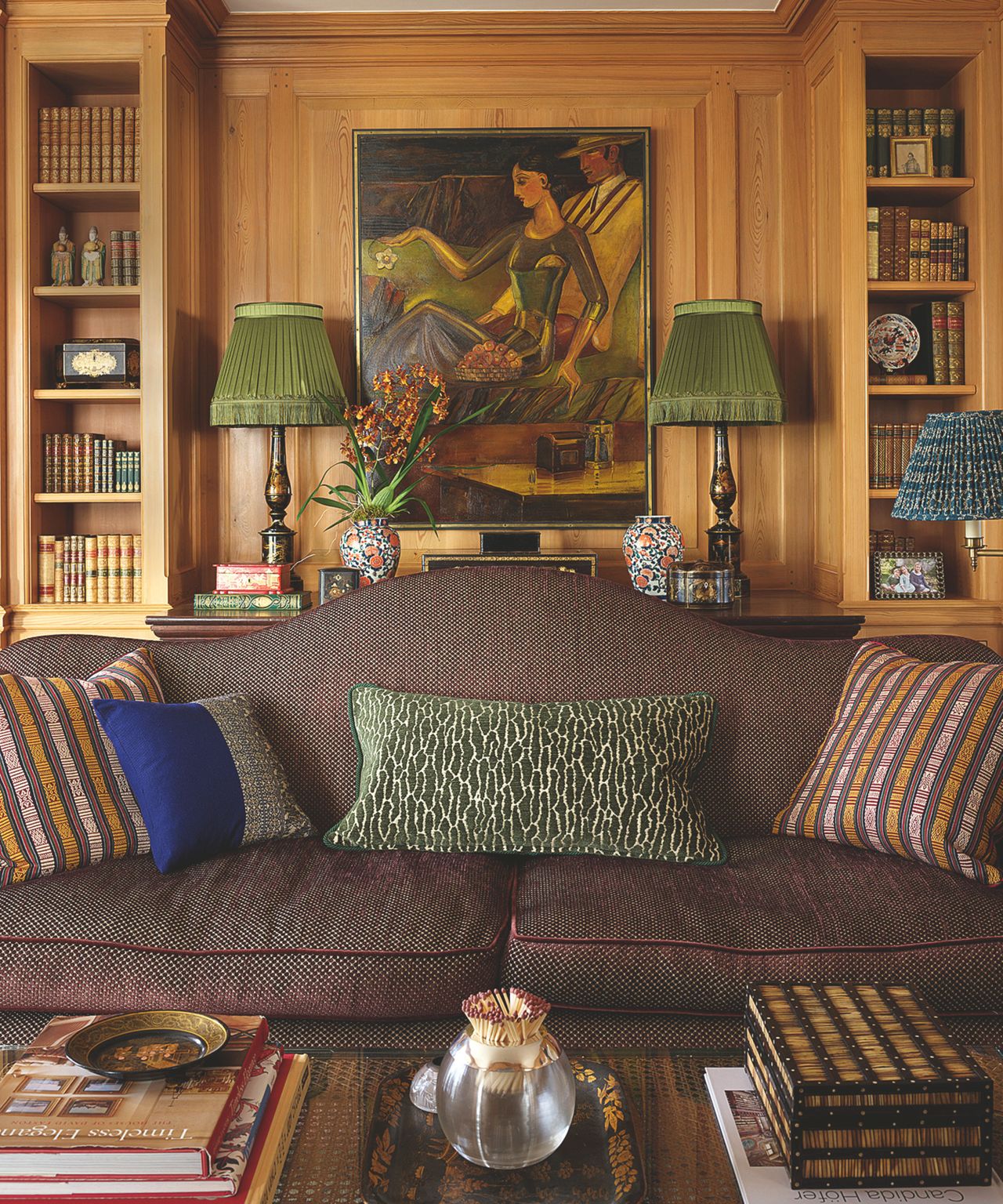 Cozy living room with light wood paneling, built-in bookshelves, a brown patterned sofa, and two lamps with pleated green shades flanking a painting