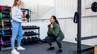 Woman performs squat holding a dumbbell by her chest in the goblet position. A trainer looks on