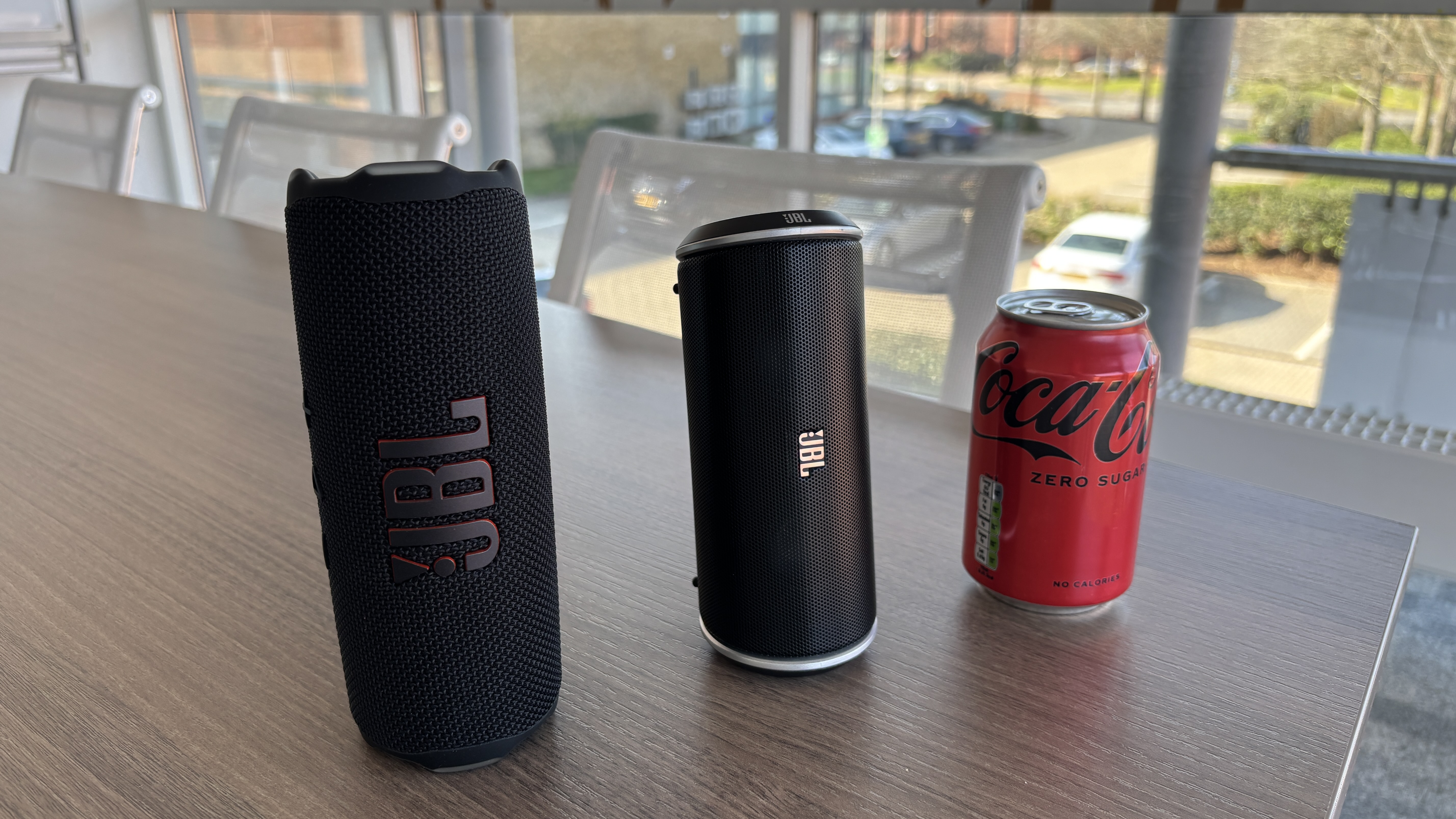 JBL Flip and Flip 7 on a desk standing upright next to a can of Coca-Cola Zero for comparison