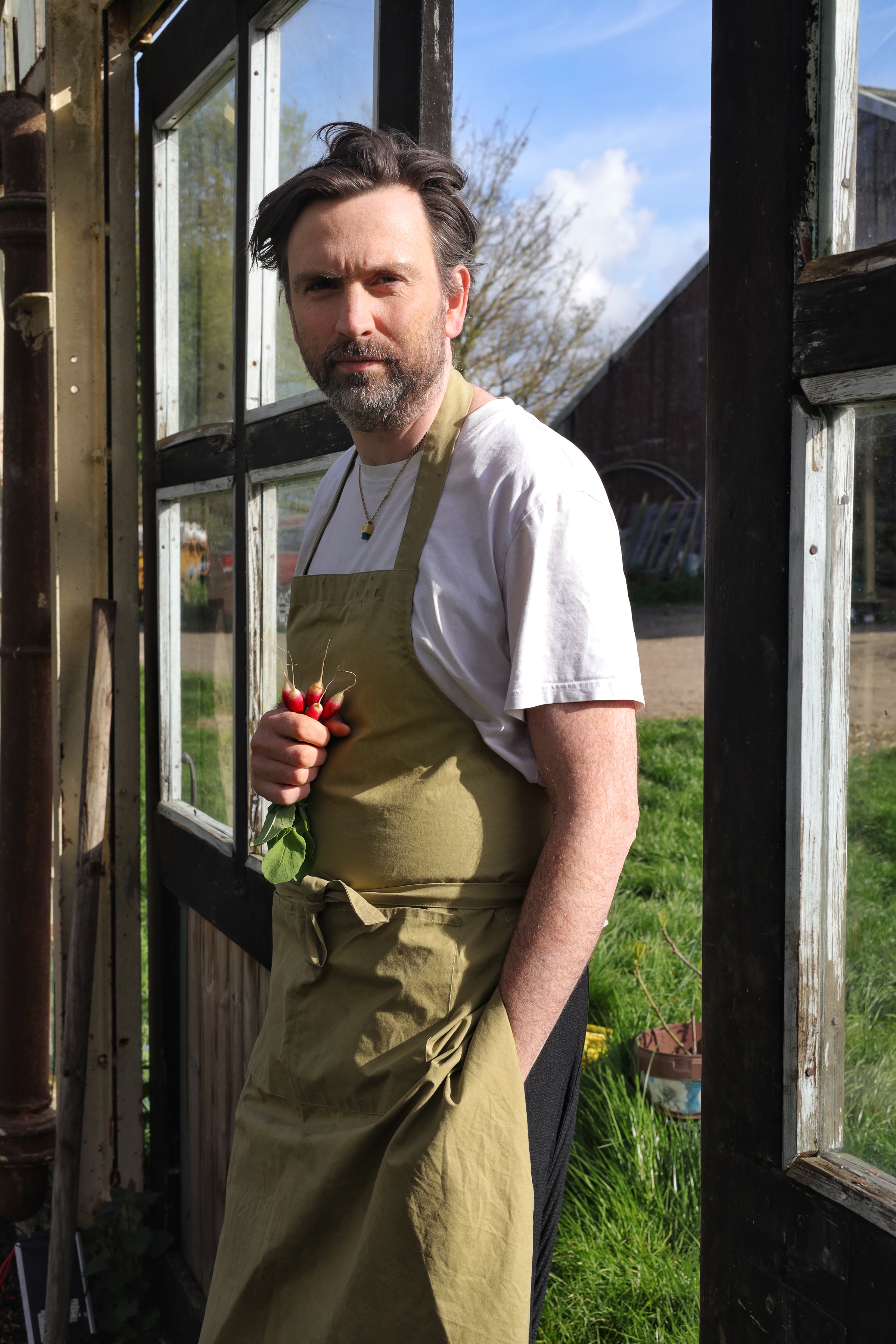 A young man in a white T-shirt and a muted lime green apron stands against a windowed wall while staring at the camera with a bunch of radish in his right hand.